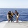 Army Divers plunge into the Pacific during exercise "Deep Blue" Two crew members in matching dark shirts and khaki shorts are assisting a scuba diver on the stern ramp of a vessel at sea. The diver is wearing a full black wetsuit with a yellow diving apparatus, and appears to be either preparing to enter or recovering from the water. The scene takes place on what appears to be a military or professional dive support vessel in open blue ocean.
