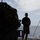 Army Divers plunge into the Pacific during exercise "Deep Blue" The image shows a silhouetted scene on the deck of a naval or military vessel at sea. In the foreground, a tactical helmet with night-vision goggle mounts is visible, held by an unseen person. In the background, a person in casual military attire stands near the ship's railing, with another vessel visible on the horizon.