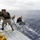 Army Divers plunge into the Pacific during exercise "Deep Blue" Two U.S. Army Deep Sea Divers in full diving gear are entering the ocean from the deck of a white vessel, while two support personnel on deck manage lines and equipment. The support personnel are wearing matching black 'U.S. Army Deep Sea Diver' t-shirts. The scene takes place in open ocean under overcast skies.