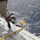 Army Divers plunge into the Pacific during exercise "Deep Blue" Two divers in full diving gear, including yellow helmets and black wetsuits, are in the water alongside a white vessel. A person on the boat deck, wearing khaki shorts and visible leg tattoos, appears to be assisting the divers. A yellow rope is visible along the boat's hull, likely used as a safety or guide line.