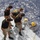 Army Divers plunge into the Pacific during exercise "Deep Blue" Three individuals on the deck of a vessel are assisting a diver who is partially in the water at the edge of the boat. The support crew, wearing matching black t-shirts with yellow graphics, are handling ropes and equipment to help the diver. The scene appears to take place in open ocean under bright sunlight.