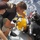 Army Divers plunge into the Pacific during exercise "Deep Blue" Military personnel, identifiable by U.S. Army apparel, are assisting a diver suited in a black wetsuit and full diving helmet equipment at the edge of a vessel. The diver is wearing a yellow hard-shell diving helmet with chrome fittings and a black scuba tank setup. The scene takes place on what appears to be a military or support vessel with open ocean visible in the background.
