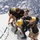 Army Divers plunge into the Pacific during exercise "Deep Blue" A group of personnel in matching black t-shirts are assisting a diver wearing a yellow hard-hat diving helmet and a rebreather/diving apparatus on the deck of a vessel. The scene appears to be a military or professional diving operation, with the team helping the diver either prepare for entry or recover from the water. The open ocean is visible in the background, suggesting an offshore operation.