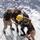 Army Divers plunge into the Pacific during exercise "Deep Blue" Three personnel in matching dark t-shirts are assisting a fully equipped deep-sea diver on what appears to be the deck ramp of a naval or dive support vessel. The diver is wearing a distinctive yellow hard-hat diving helmet, a dry suit, and a back-mounted air supply system. The scene takes place at sea, with open blue ocean water visible in the background.