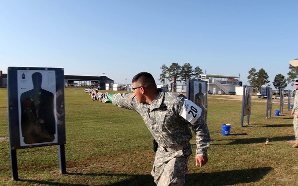 A military shooting range exercise is underway outdoors, with a soldier in ACU (Army Combat Uniform) wearing bib number 20 aiming a pistol at a silhouette target. A civilian instructor or observer in a black shirt and tan cap stands nearby watching. Additional soldiers and targets are visible in the background across a grassy field.