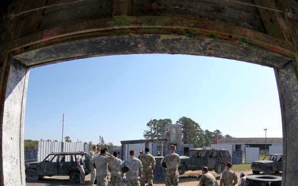 The image is taken through a window opening using a fisheye lens, showing a group of military personnel in camouflage uniforms gathered in an outdoor training area. The scene includes burned-out vehicles, metal barrels, stacked tires, and modular buildings, consistent with a military urban operations or combat training facility. The fisheye distortion creates a curved, barrel-shaped perspective of the exterior scene.