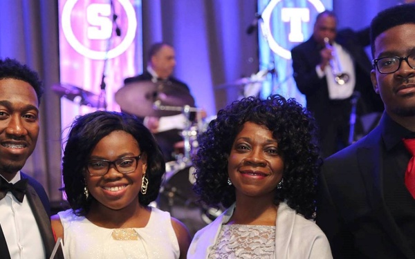 Four individuals are posing together at what appears to be a formal awards gala or banquet event. The person on the far left is holding a glass/crystal award trophy. A live band is visible performing in the background.
