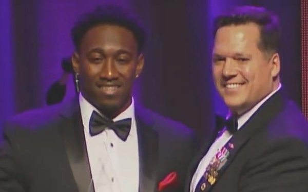 Two men in formal attire are on a stage sharing a crystal/glass award trophy. One man is wearing a tuxedo with a bow tie and red pocket square, while the other appears to be a military officer wearing a dark dress uniform with gold sleeve stripes, medals, and ribbons. The setting appears to be a formal awards ceremony or gala event.