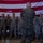Lt. Gen. Nicholson recognizes Airmen’s support in MV-22 incident A group of military personnel are gathered in formation inside what appears to be a hangar or large indoor facility, with a large American flag displayed on the wall behind them. One individual in Marine Corps MARPAT woodland camouflage is facing the group with their back to the camera, appearing to address or brief the assembled airmen. The majority of personnel are wearing Air Force Airman Battle Uniform (ABU) tiger-stripe camouflage pattern.