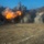 KMEP 17-8 The image shows a large explosion occurring in an arid, open terrain area near a hillside or rocky embankment. Bright orange fireballs are visible at the center of the blast, surrounded by billowing black and gray smoke clouds rising into a clear blue sky. The foreground shows dry, rocky ground with scattered debris and dust.