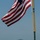 31 MEU offloads equipment off of USS Green Bay in support of Exercise Gold Gold 2017 A large American flag flies prominently from a flagpole on what appears to be the deck of a naval vessel. In the background, a smaller gray military vessel is visible on the water, with hilly or mountainous terrain visible on the horizon. The scene suggests a naval operation or port visit in a coastal area.