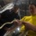170213-N-WV703-025 Two individuals are handling a large snake in what appears to be an industrial or military setting. The man in the foreground wearing a yellow long-sleeve shirt with a visible tattoo on his arm is holding the snake's head, while the person in the background wearing a dark shirt and blue cap assists. The environment behind them appears to be a metal-walled space with pipes or equipment visible.