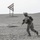 FASTCENT conduct live-fire training in CENTCOM AOR A soldier in full combat gear runs across a sandy desert terrain. The individual is wearing digital camouflage (MARPAT pattern consistent with U.S. Marines), helmet with mounted device, ballistic goggles, and carries a rifle and backpack. In the background, a triangular metal range target structure is visible on a sand dune.