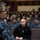 170215-N-JD834-014 A military briefing or all-hands meeting is taking place in an auditorium filled with U.S. Navy personnel in blue digital camouflage (NWU Type I) uniforms. A speaker, visible from behind in the foreground, addresses a large seated audience of sailors. The setting appears to be a formal military assembly hall with tiered seating.