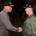 Capt. Lenox welcomes aboard Rear Adm. Kreitz Two military personnel in U.S. Navy uniforms are shaking hands in what appears to be the hangar bay of an aircraft carrier. The individual on the left wears a gray flight suit with a command cap bearing gold insignia, while the individual on the right wears an olive green flight jacket with a name/wing badge and a navy blue command cap. Additional uniformed personnel are visible in the background.