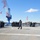 170214-N-YG414-100 A military formation is assembled on the flight deck of a U.S. Navy vessel, with personnel wearing blue camouflage uniforms and blue hard hats. A single officer in a white hard hat appears to be addressing the assembled crew via a microphone. The ship is docked at a port facility, visible by the large red and blue cranes in the background, with an American flag flying on deck.