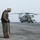 Group Sails A military deck crew member stands on what appears to be a naval vessel's flight deck, holding tie-down chains used to secure aircraft. In the background, a large military helicopter, identifiable as a CH-53E Super Stallion, is parked on the deck. The scene suggests flight deck operations aboard an amphibious assault ship under overcast conditions.