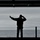 Sailor tests elevator A silhouetted figure stands on what appears to be the deck of a naval vessel, facing the open ocean under an overcast sky. The person is gesturing with one arm extended and pointing while holding an object, likely a radio or communication device, to their face. The image is a strong backlit silhouette composition with the sea and grey sky as the backdrop.