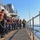 170214-N-NJ910-0037 A group of naval personnel are lined up along the exterior deck of a military vessel, handling a rope or line in what appears to be a seamanship drill or mooring evolution. Most crew members are wearing orange life vests over dark navy uniforms, while two supervisory figures at the far end wear yellow vests and a white hard hat. The scene takes place on calm open water under clear blue skies.