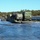 Bridge Company crosses the gap A military ferry or pontoon raft is transporting multiple military trucks across a body of water, likely during a training exercise. The vehicles are loaded with covered cargo and display orange hazard diamond placards. A smaller support vessel is visible to the left, and the operation takes place near a wooded shoreline under clear skies.