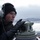 170207-N-HI376-022 A young man wearing a black knit cap, communication headset with boom microphone, and black leather gloves is operating a nautical bearing compass or pelorus mounted on what appears to be the bridge wing of a vessel. The setting is a cold, wintry maritime environment with icy water, overcast skies, and snow-dusted mountains visible in the background. His focused expression and equipment suggest he is performing navigational duties aboard a ship.