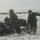In the CRRC of Time: U.S. Marines and Japanese Soldiers conduct Recon Drills at Exercise Iron Fist Military personnel in camouflage uniforms are conducting a beach landing or surf passage exercise with a black inflatable rubber raiding craft (CRRC) in rough surf conditions. At least three individuals are visible managing the craft at the shoreline, with one person appearing to give directional signals. The overcast conditions and choppy surf suggest a realistic training environment.