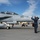 170206-N-IM832-013 A U.S. Navy aircraft handler in digital camouflage uniform is marshaling a twin-seat F/A-18 Hornet (bureau number 105) on a military airfield tarmac. The handler is giving a thumbs-up signal while wearing a red helmet/cranial protector. The scene appears to be a routine flight operations evolution at a naval air station.