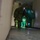 Chemical, Biological, Radiological and Nuclear Marines Participate Advanced Training Personnel in hazmat/CBRN protective suits are conducting what appears to be a training exercise inside a building corridor. At least three individuals are visible wearing different colored encapsulated suits (olive/dark green and bright green), with labels reading 'TRAINING SUIT ONLY' visible on the suits. The low-angle perspective from a doorway shows an illuminated hallway with an EXIT sign overhead.
