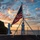 170211-N-BL637-021 Two U.S. Navy sailors in dress white uniforms are raising the American flag on what appears to be the deck of a naval vessel at sunrise or sunset. The scene is set against a dramatic sky with orange and blue clouds, with a coastline and ocean visible in the background. The ceremonial flag-raising is conducted using a portable tripod flagpole mounted on the ship's deck.