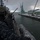 170207-N-HI376-002 Two naval personnel in combat helmets and digital camouflage uniforms stand watch on the deck of a warship as it approaches or departs a snow-covered port facility. The scene appears to be taken during winter conditions with visible snowfall, and green industrial cranes are visible on the pier in the background. The port infrastructure, including warehouses and vehicles, suggests a military or commercial harbor in a cold-weather region.