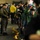 Nimitz Sailors undergo inspection A military or naval personnel member in an olive/flight suit is distributing or demonstrating equipment to a line of young people, likely Boy Scouts or cadets, in what appears to be a hangar bay or large indoor military facility. The young visitors are holding helmets and gear with attached cords and orange tags, suggesting a safety equipment demonstration or orientation. Several crew members in yellow shirts and Navy digital camouflage uniforms are also present, indicating this may be a ship or base tour event.