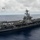 170209-N-GD109-180 A large U.S. Navy aircraft carrier bearing hull number '70' is underway in open ocean under partly cloudy skies. The flight deck is densely populated with numerous fixed-wing aircraft and helicopters. A single aircraft, appearing to be a turboprop type, is airborne above the ship.