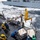 170212-N-HI376-194 A Replenishment at Sea (RAS/UNREP) operation is underway between two naval vessels in rough ocean conditions. Sailors wearing color-coded safety helmets and life vests are guiding palletized cargo being transferred via a tensioned highline and crane system. The green vests marked 'UNREP' and 'UNREP TEAM' indicate this is a coordinated underway replenishment evolution.