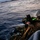 RIMPAC’s lost shots Two military personnel in camouflage uniforms and tactical gear are positioned on what appears to be a small watercraft at sea during low-light conditions, likely dusk or dawn. They are equipped with helmets, body armor, and tactical vests, and appear to be on a maritime patrol or insertion operation. A rocky landmass or island is faintly visible in the background.