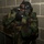 MCESG Gas Chamber A military personnel wearing woodland camouflage BDU (Battle Dress Uniform) is donning or adjusting a black gas mask/protective mask against a concrete block wall. The individual appears to be in an indoor training facility, likely participating in CBRN (Chemical, Biological, Radiological, Nuclear) defense training. A second person in similar camouflage uniform is partially visible on the left edge of the frame.