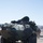 Marines and Soldiers train with RFID and Shout nano in tracking Railops A front-facing view of a Light Armored Vehicle (LAV), specifically appearing to be an LAV-AT (Anti-Tank) variant equipped with a TOW missile launcher system mounted on the turret. The vehicle is painted in a green camouflage pattern and is stationary on a paved surface in an arid, desert-like environment. At least one crew member is visible in the open hatch, and a soldier in desert camouflage uniform is walking behind the vehicle.