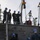 170215-N-KP948-038 A group of U.S. Navy sailors wearing blue hard hats and Navy Working Uniform (NWU) camouflage are conducting a weapons or ordnance handling evolution aboard a naval vessel. Personnel are positioned both on an upper deck and a lower deck, coordinating the movement of a cylindrical object — likely a torpedo or similar ordnance — using a chain hoist/crane system. The operation appears to be a loading or transfer exercise at a pier or alongside another vessel.