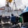 170212-N-HI376-158 Naval personnel are conducting an underway replenishment (UNREP) or cargo transfer operation on the deck of a military vessel. Workers wearing blue and green helmets are guiding palletized cargo suspended by crane rigging from an adjacent supply ship visible in the background. The operation appears to be a coordinated logistics transfer between two naval vessels.