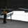 Sailor tests elevator A naval deck crew member wearing a yellow safety vest is standing on what appears to be an aircraft carrier hangar deck or flight deck edge, using hand signals with both arms extended. The person is facing away from the camera toward open water, likely directing aircraft or vessel movement. The scene is backlit, creating a strong silhouette effect against the bright water visible through the deck opening.