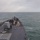 170207-N-HI376-031 The image shows the forward deck of a U.S. Navy guided-missile destroyer underway at sea in overcast, choppy conditions. Multiple sailors are visible on the bow deck engaged in what appears to be line-handling or seamanship evolutions. A 5-inch Mk 45 naval gun mount is prominently visible on the forecastle, along with vertical launch system (VLS) cells in the foreground.