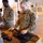 170215-N-N0901-001 U.S. Navy personnel in NWU (Navy Working Uniform) digital camouflage are conducting weapons familiarization or maintenance training at a table. Multiple service members are handling disassembled M4/M16-style rifles laid out on a padded surface. The setting appears to be an indoor training facility or gymnasium.