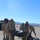 Marines and Soldiers train with RFID and Shout nano in tracking Railops A group of military personnel in desert camouflage uniforms are handling large dark-colored duffel bags or equipment bundles on a paved surface in an arid desert environment. Two soldiers are actively carrying or repositioning a heavy bag while others observe nearby. The setting appears to be a military installation or airfield with mountains visible in the background.