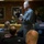 INSURV President adresses Nimitz A senior U.S. Navy officer in a gray flight suit is addressing a group of personnel seated at tables in what appears to be a briefing or dining room. Multiple attendees are wearing jackets with 'INSURV' patches and USS Nimitz (CVN-68) insignia on the backs of their chairs. A framed portrait of a naval officer hangs on the brick wall in the background.