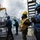 170212-N-HI376-271 A replenishment-at-sea (RAS) operation is underway on the deck of a U.S. Navy vessel, with sailors in various colored safety vests and helmets managing cargo pallets being transferred via crane rigging. At least four personnel are visible working to secure and guide a pallet of supplies, with a second ship visible in the background. The overcast conditions and cold-weather gear suggest operations in a northern or winter environment.