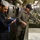 Inspector corrects discrepancies Two U.S. Navy personnel are conducting what appears to be a galley inspection aboard a naval vessel. The individual on the right, identified by name tape as 'GIBSON,' is wearing gray coveralls with officer insignia and a command ball cap, and is inspecting a stainless steel sink area. The individual on the left is in navy blue coveralls with a white cook's cap and is recording notes on a clipboard.