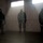 MCESG Gas Chamber Three individuals wearing military camouflage uniforms and gas masks are standing inside a concrete block building filled with haze or smoke. Light streams in from two high windows, illuminating the smoky atmosphere. This appears to be a military CBRN (Chemical, Biological, Radiological, Nuclear) training exercise conducted in a gas chamber facility.