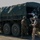 Wanted: Dead or Alive Military personnel in combat gear are dismounting or loading onto a large olive-drab military cargo truck, likely during a training exercise. Several soldiers wearing camouflage uniforms, helmets, and tactical vests are visible around the vehicle on a dirt road. The setting appears to be a training range or field exercise area with green hills in the background.