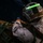 RIMPAC’s lost shots A military service member wearing desert MARPAT camouflage and a tactical helmet is photographed in low-light conditions. A bright green chemlight (glow stick) is attached horizontally to the front of the helmet, serving as a visual identification marker. The individual appears to be handling or securing a large pack or equipment bag.
