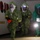 Chemical, Biological, Radiological and Nuclear Marines Participate Advanced Training Three individuals wearing full hazmat/CBRN protective suits are moving through what appears to be a dimly lit indoor corridor or building. Two are wearing olive/green encapsulated suits with reflective face shields, while a third wears a teal/turquoise suit labeled 'TRAINING SUIT ONLY'. The setting appears to be a military or emergency response training exercise, indicated by the 'MCJR' lettering and 'UNSELFISHNESS' text visible on the wall.