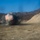 KMEP 17-8 An explosion is captured mid-detonation in an open, arid valley or quarry-like terrain surrounded by hills with sparse, dry vegetation. The blast produces a large cloud of black and orange-tinged smoke rising from the ground. The image appears to document a controlled demolition or ordnance disposal event based on the isolated, prepared setting.
